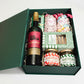 A green magnetic box containing a variety of Christmas treats including wine, chocolates, crackers, and condiments.
